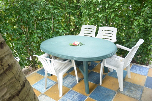 Table ronde en plastique vert avec quatre chaises blanches, entourée de verdure, sur un sol en carreaux colorés.